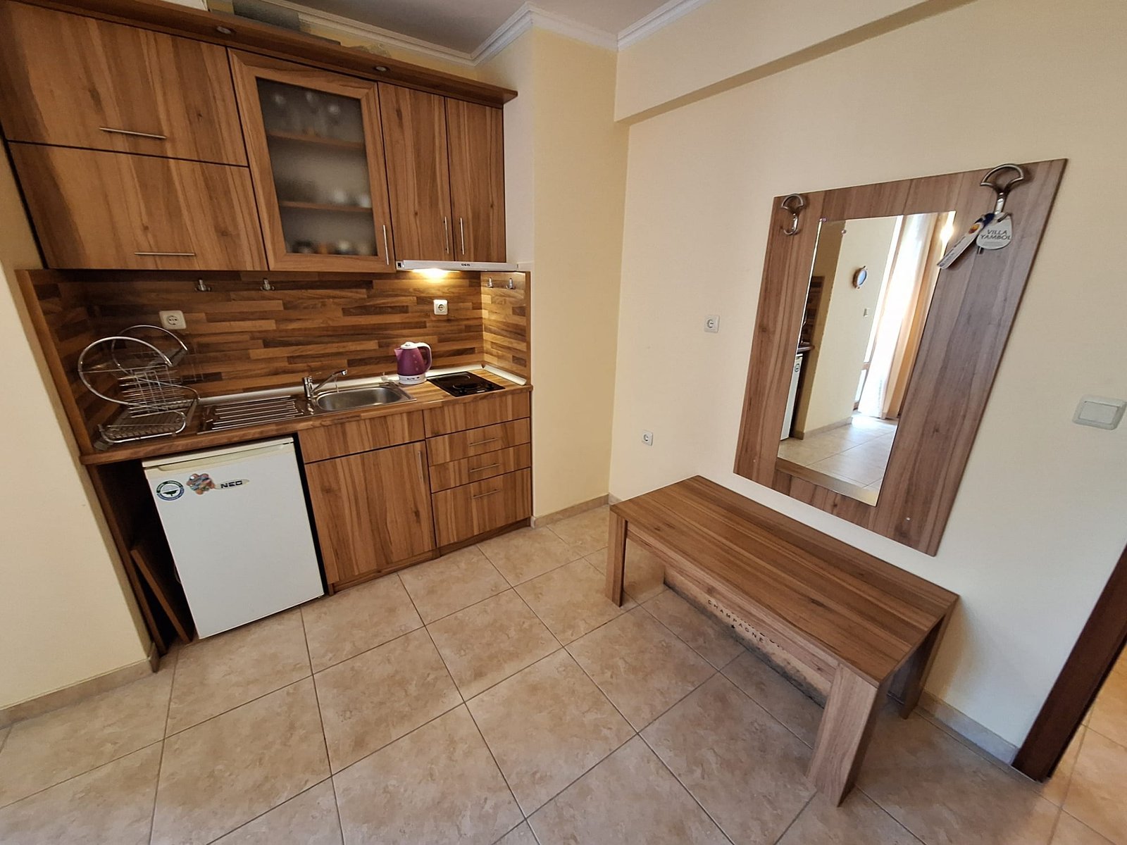 Современная кухня с деревянными шкафами, мойкой, плитой и зеркалом, рядом деревянная скамейка в светлом интерьере.