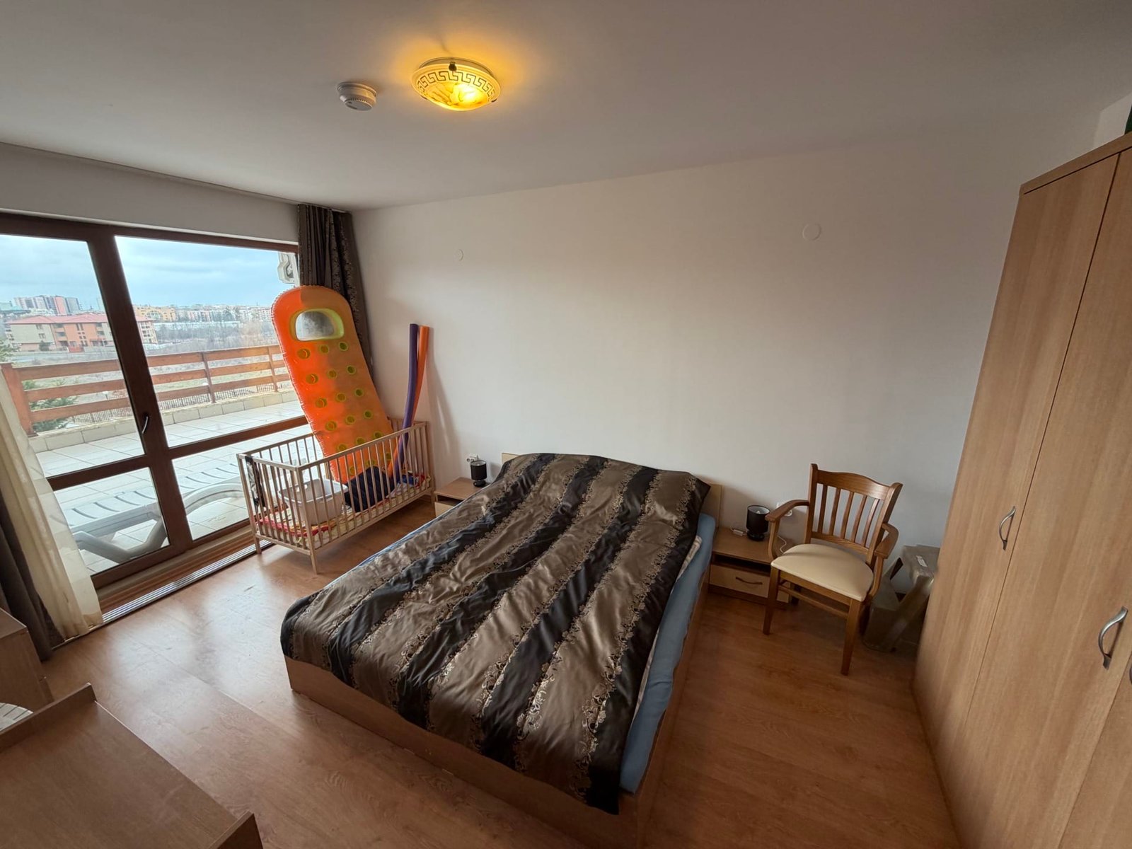 Современная спальня с двуспальной кроватью, комодом, креслом и детской кроваткой, выход на балкон с панорамным видом.