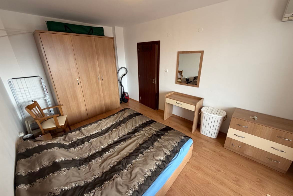 Комната с двуспальной кроватью, деревянной мебелью, зеркалом и шкафом, светлые стены и пол, аккуратный интерьер.