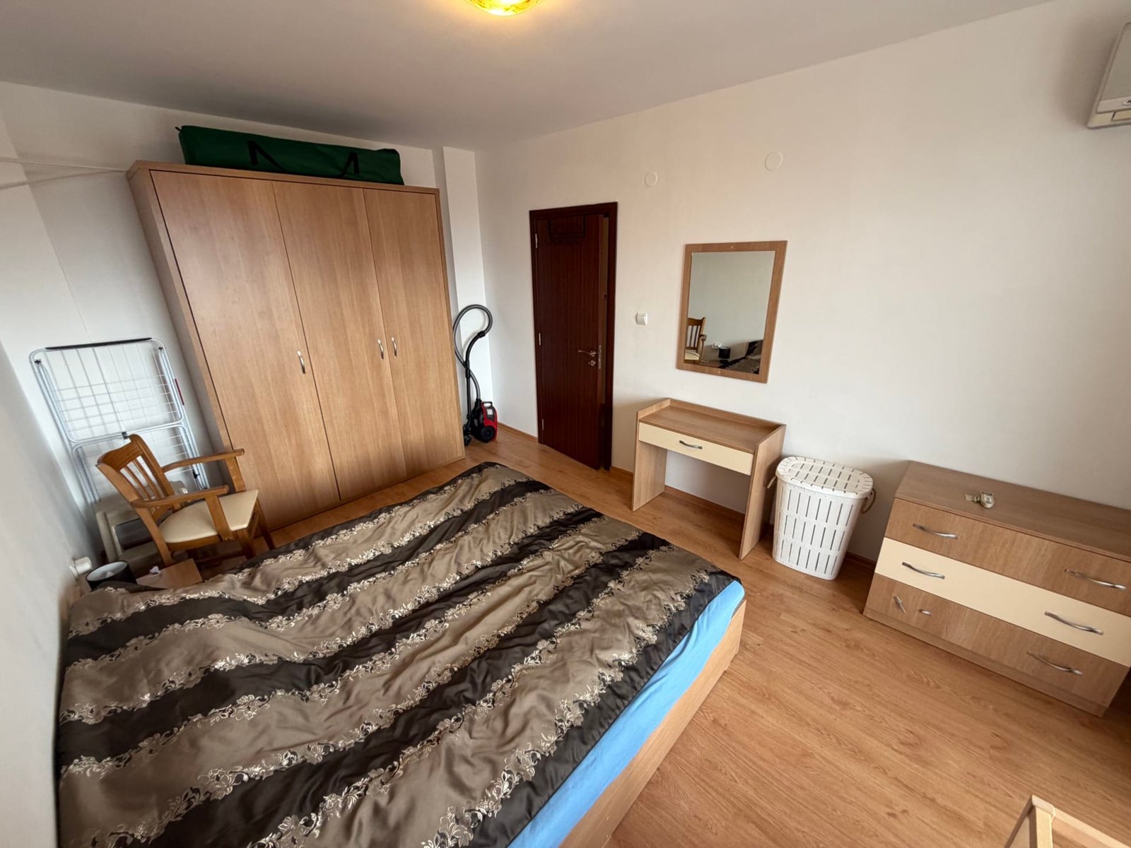 Комната с двуспальной кроватью, деревянной мебелью, зеркалом и шкафом, светлые стены и пол, аккуратный интерьер.