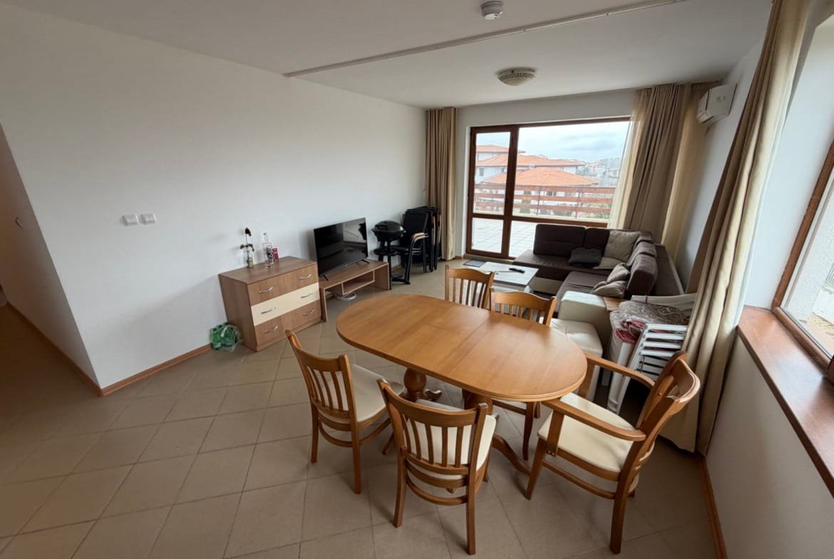 Светлая гостиная с обеденным столом, стульями, диваном и телевизором, окна с видом на окружающий ландшафт.