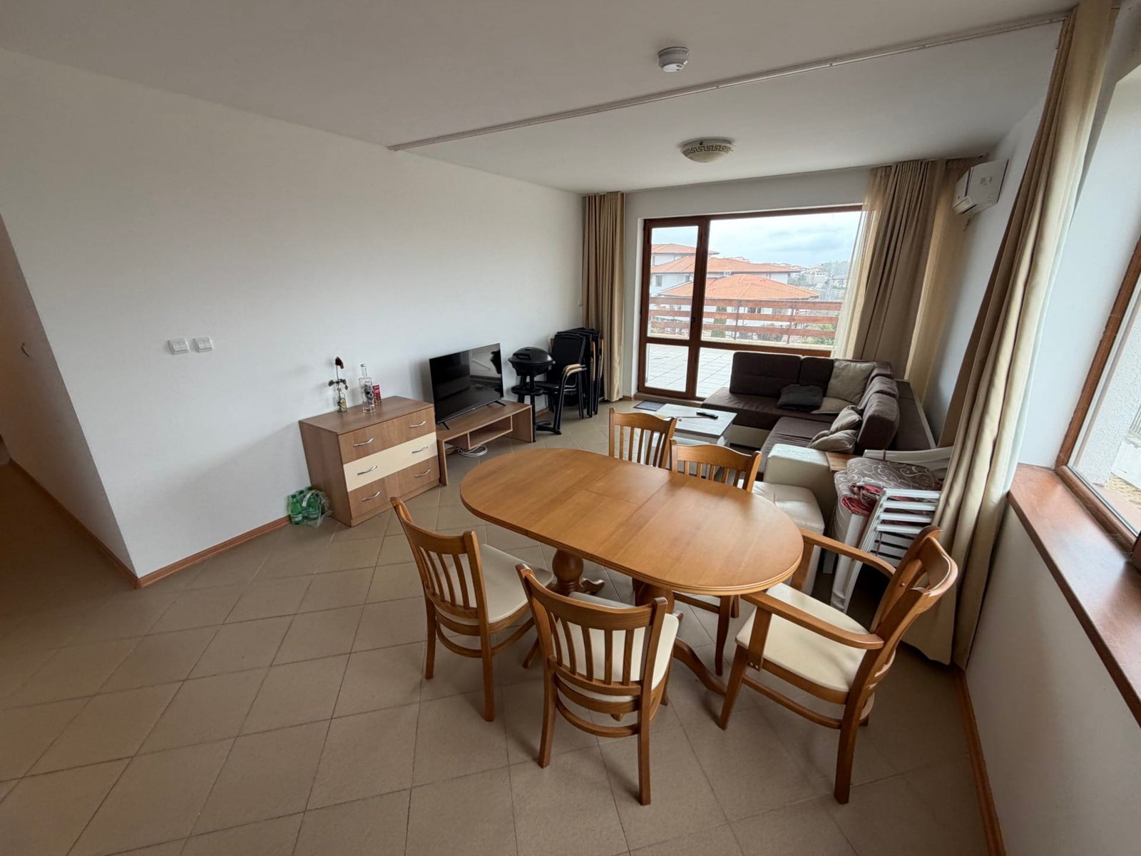 Светлая гостиная с обеденным столом, стульями, диваном и телевизором, окна с видом на окружающий ландшафт.