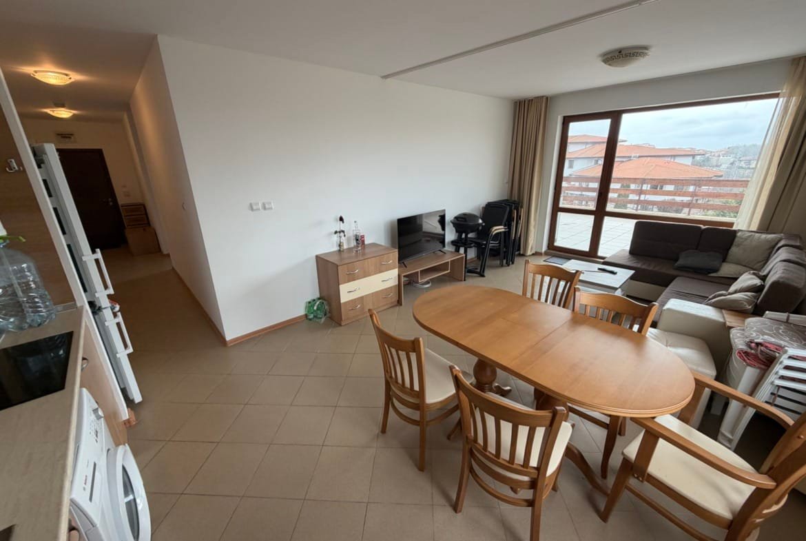 Современный трёхкомнатный апартамент с обеденной зоной, диваном и телевизором, вид на балкон и интерьер в светлых тонах.