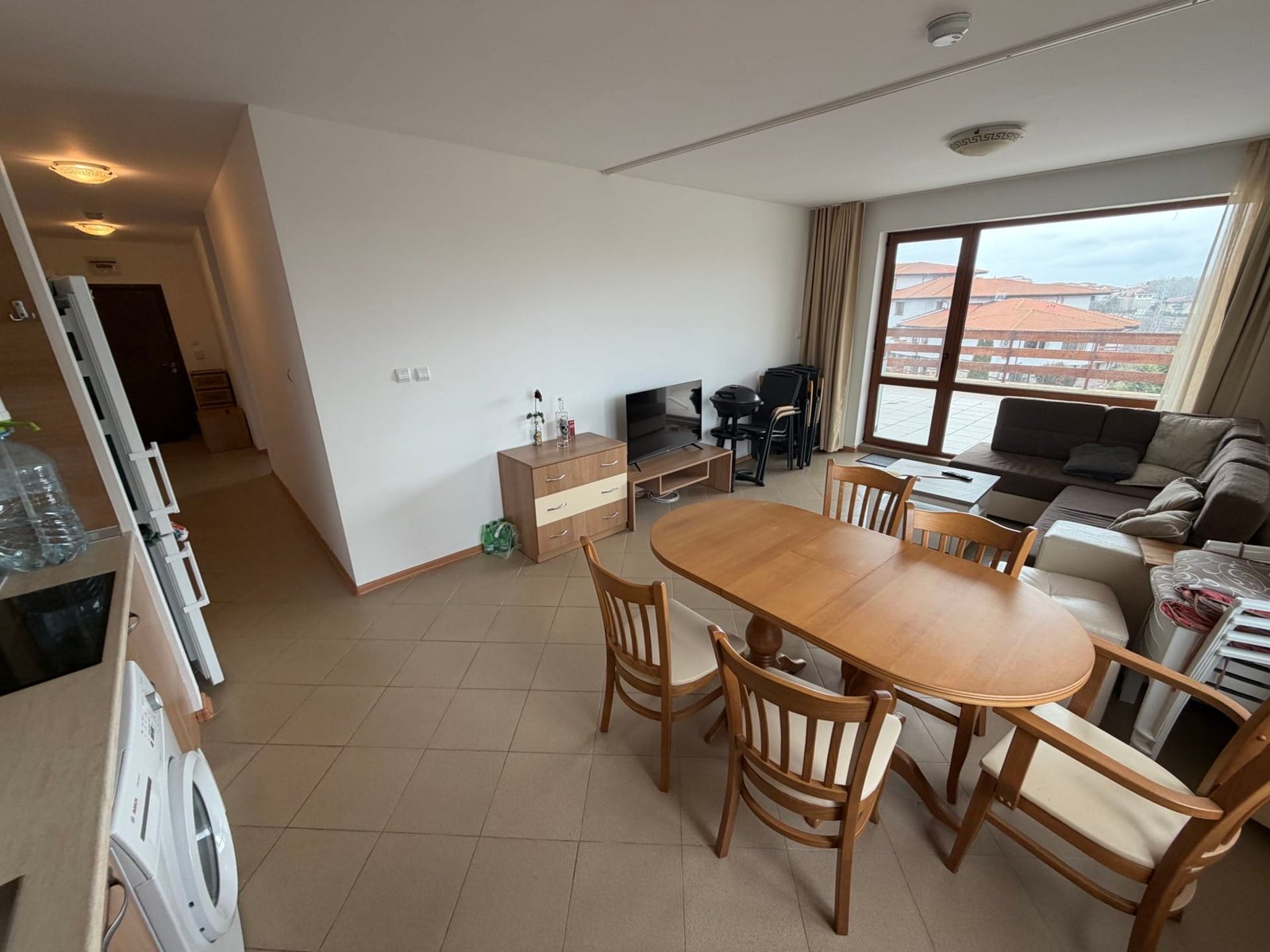 Современный трёхкомнатный апартамент с обеденной зоной, диваном и телевизором, вид на балкон и интерьер в светлых тонах.