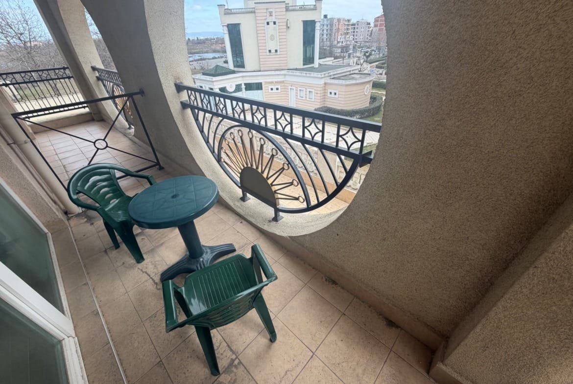 Балкон двухкомнатного апартамента с круглыми перегородками и столом, зелеными стульями, видом на окружающие здания.