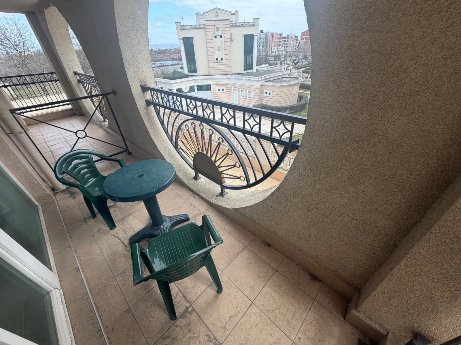 Балкон двухкомнатного апартамента с круглыми перегородками и столом, зелеными стульями, видом на окружающие здания.