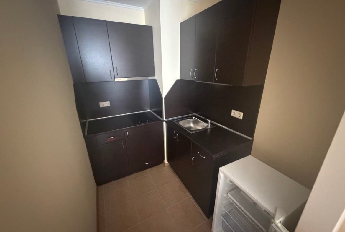 Кухонный уголок в двухкомнатном апартаменте: темные шкафчики, раковина и холодильник на светлом полу.