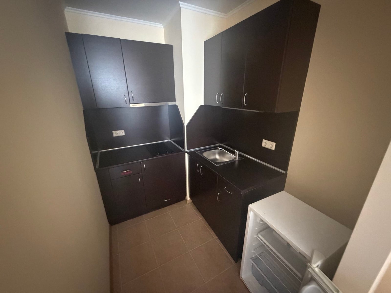 Кухонный уголок в двухкомнатном апартаменте: темные шкафчики, раковина и холодильник на светлом полу.