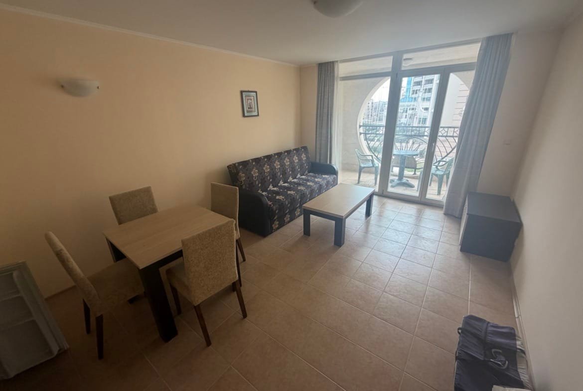 Двухкомнатный апартамент с диваном, столом и стульями, светлые стены и большие окна с видом на балкон.