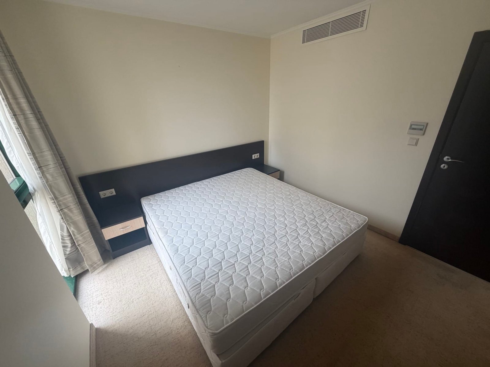 Двуспальная кровать без матраса в светлом комнате с окнами и нейтральными тонами, современный интерьер.