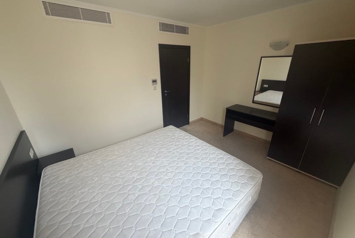 Современная спальня с кроватью без матраса, темной мебелью и зеркалом на стене, светлые стены и пол.