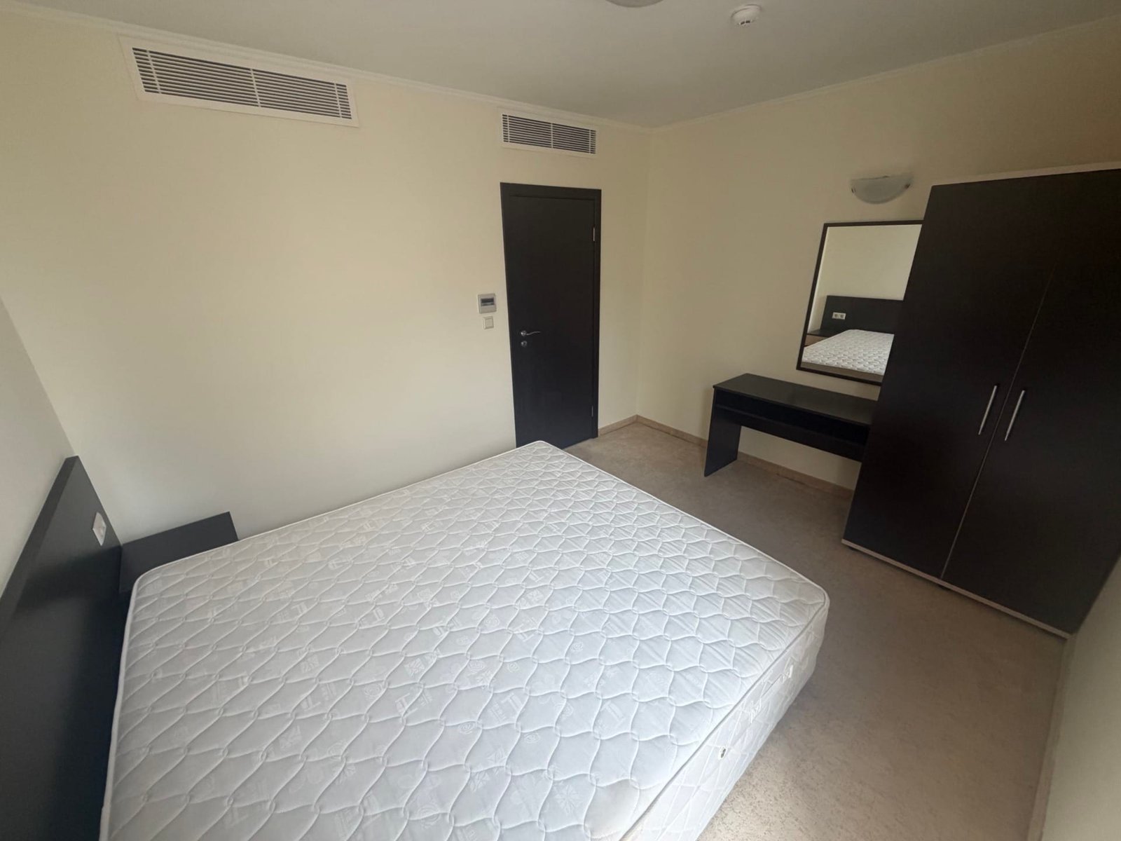 Современная спальня с кроватью без матраса, темной мебелью и зеркалом на стене, светлые стены и пол.