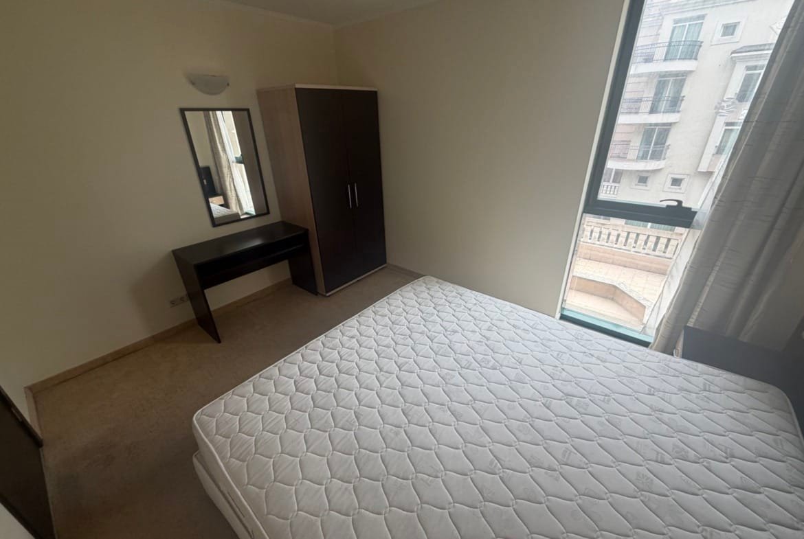 Двухкомнатный апартамент с кроватью, зеркалом и шкафом, окна с видом на балкон и светлые стены.