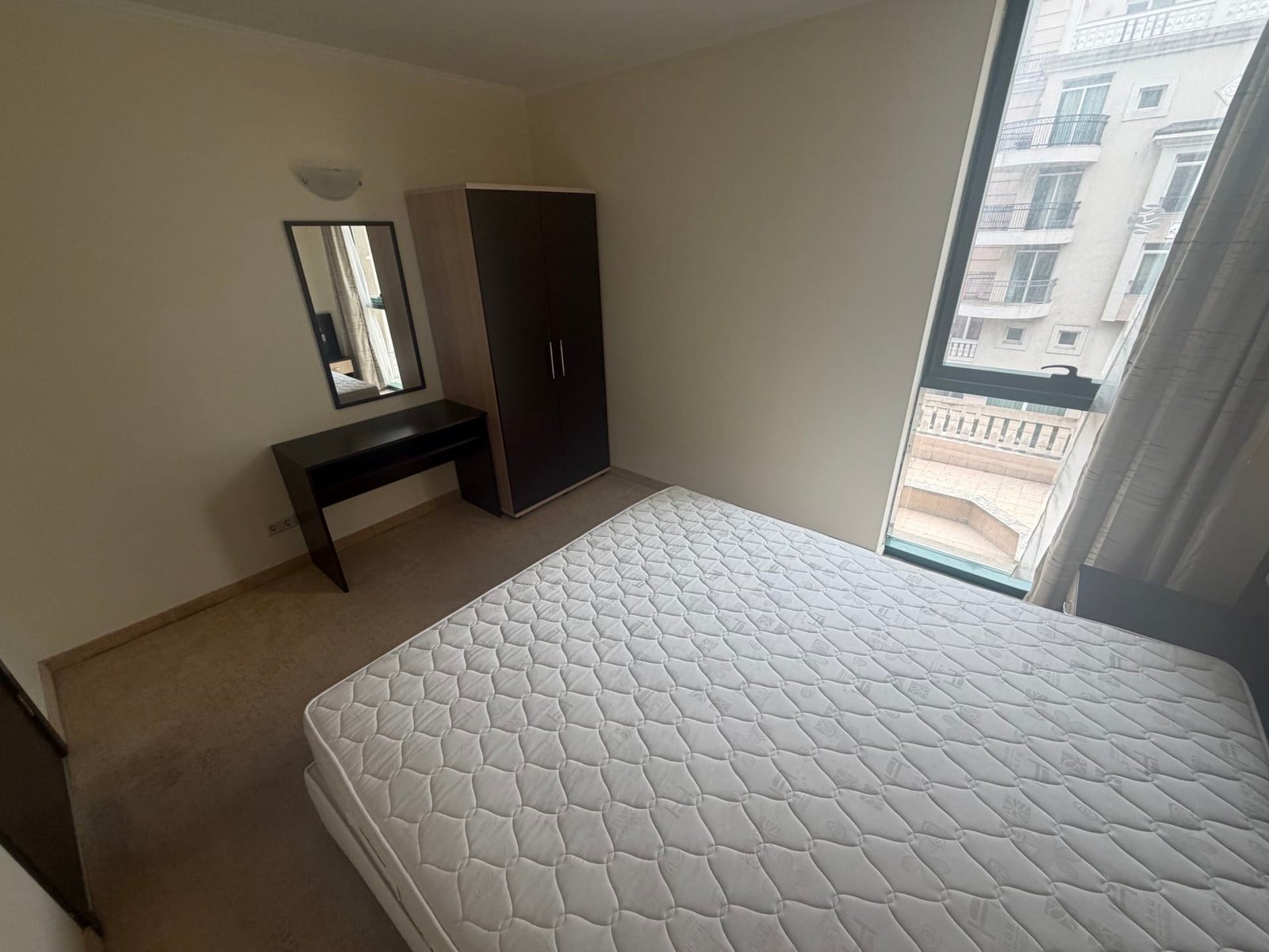 Двухкомнатный апартамент с кроватью, зеркалом и шкафом, окна с видом на балкон и светлые стены.