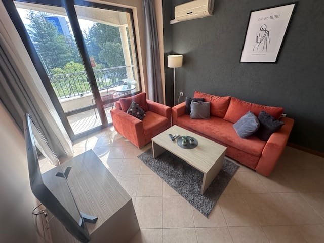 Светлая гостиная с оранжевым диваном, креслом и телевизором, выходящая на балкон с зелеными деревьями вне.