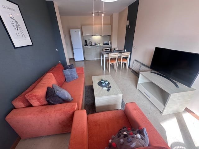 Светлая гостиная с оранжевым диваном, телевизором и кухонной зоной, оформленная в современном стиле.