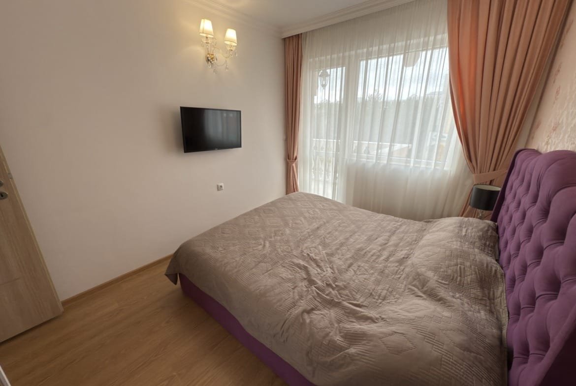 Современная спальня с серо-розовым текстильным оформлением, телевизором на стене и ярким натуральным светом.