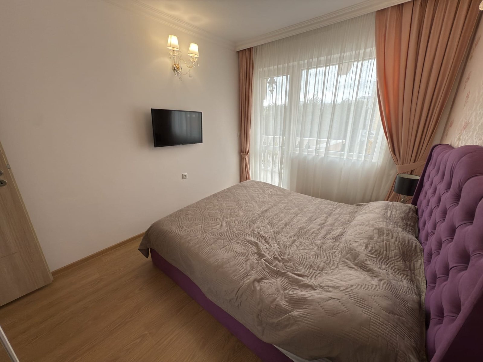 Современная спальня с серо-розовым текстильным оформлением, телевизором на стене и ярким натуральным светом.