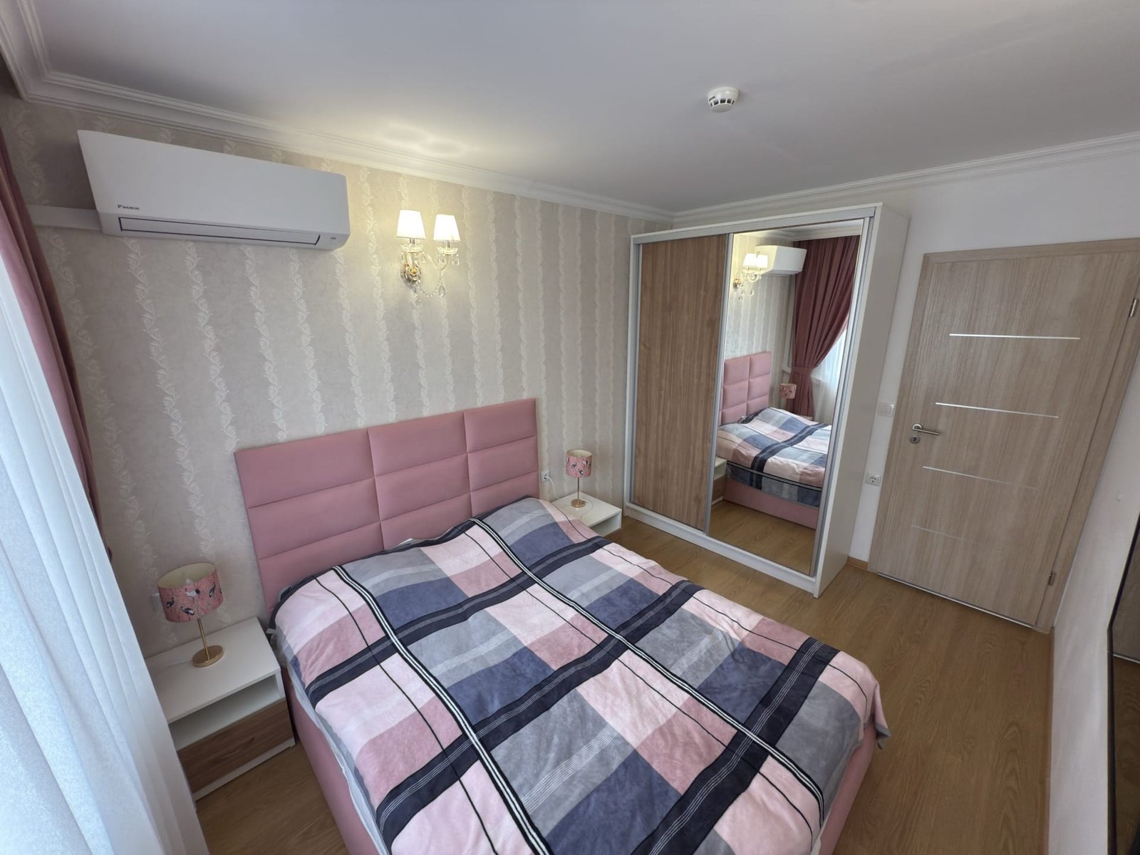 Современная спальня с двуспальной кроватью, мягкими тонами, зеркальным шкафом и светлыми шторами. Комфортное оформление.