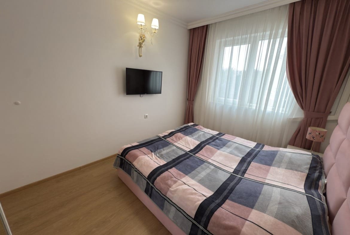 Современная спальня с кроватью, телевизором и светлыми шторами, оформленная в пастельных тонах и стильном дизайне.