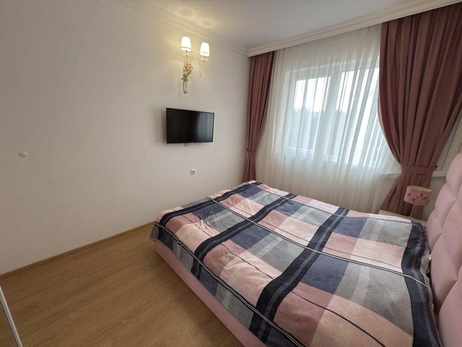 Современная спальня с кроватью, телевизором и светлыми шторами, оформленная в пастельных тонах и стильном дизайне.