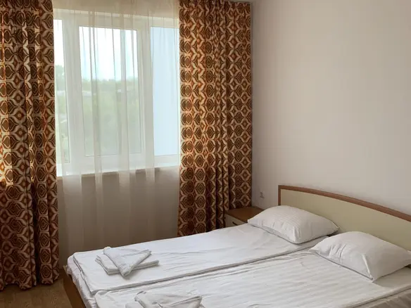 Двуспальная кровать с белыми простынями, настенные занавески с оранжевым узором, светлый интерьер, окно с видом на природу.