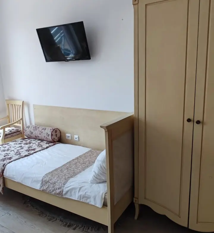 Светлая студия с отдельной кроватью, деревянным шкафом и телевизором на стене. Уютная обстановка и просторное пространство.