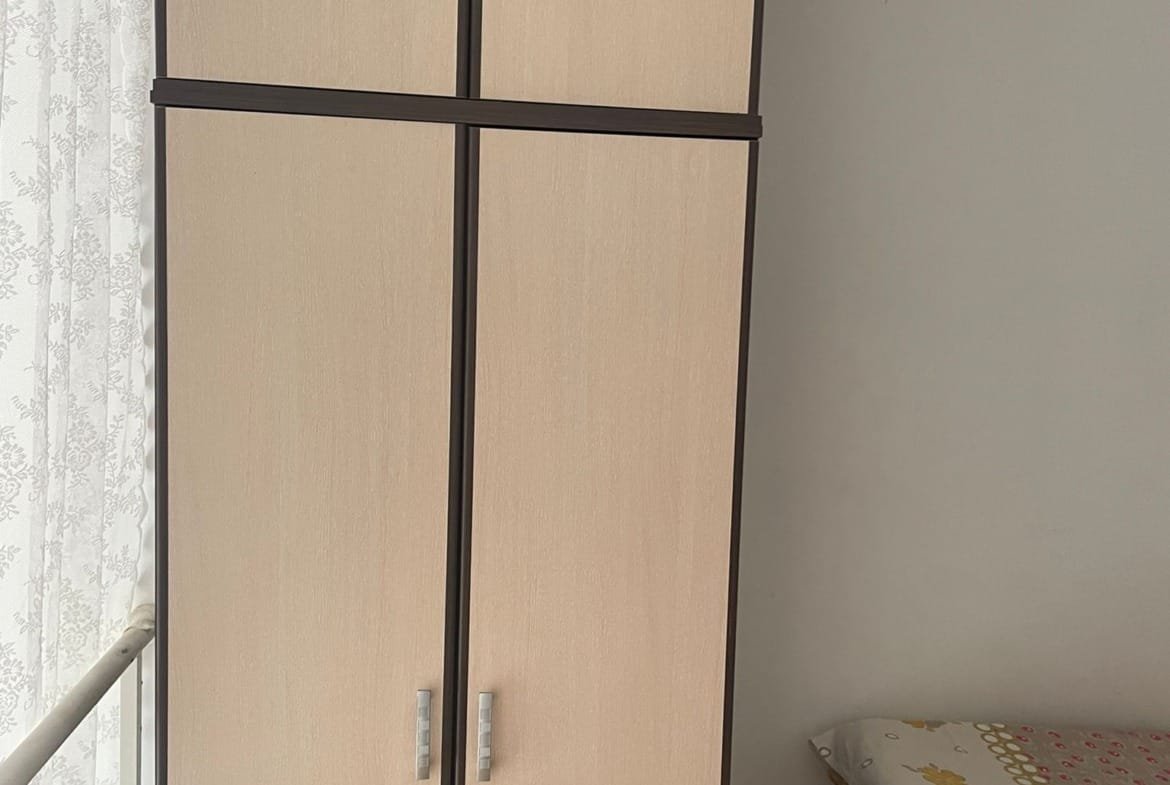 Деревянный шкаф светлого цвета с четырьмя дверцами рядом с кроватью и окнами с занавесками.