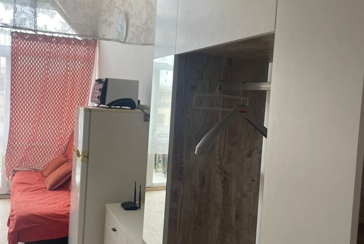 Студия с жилой зоной, диваном, холодильником и шкафом, оформленная в светлых тонах с акцентами на текстиле.