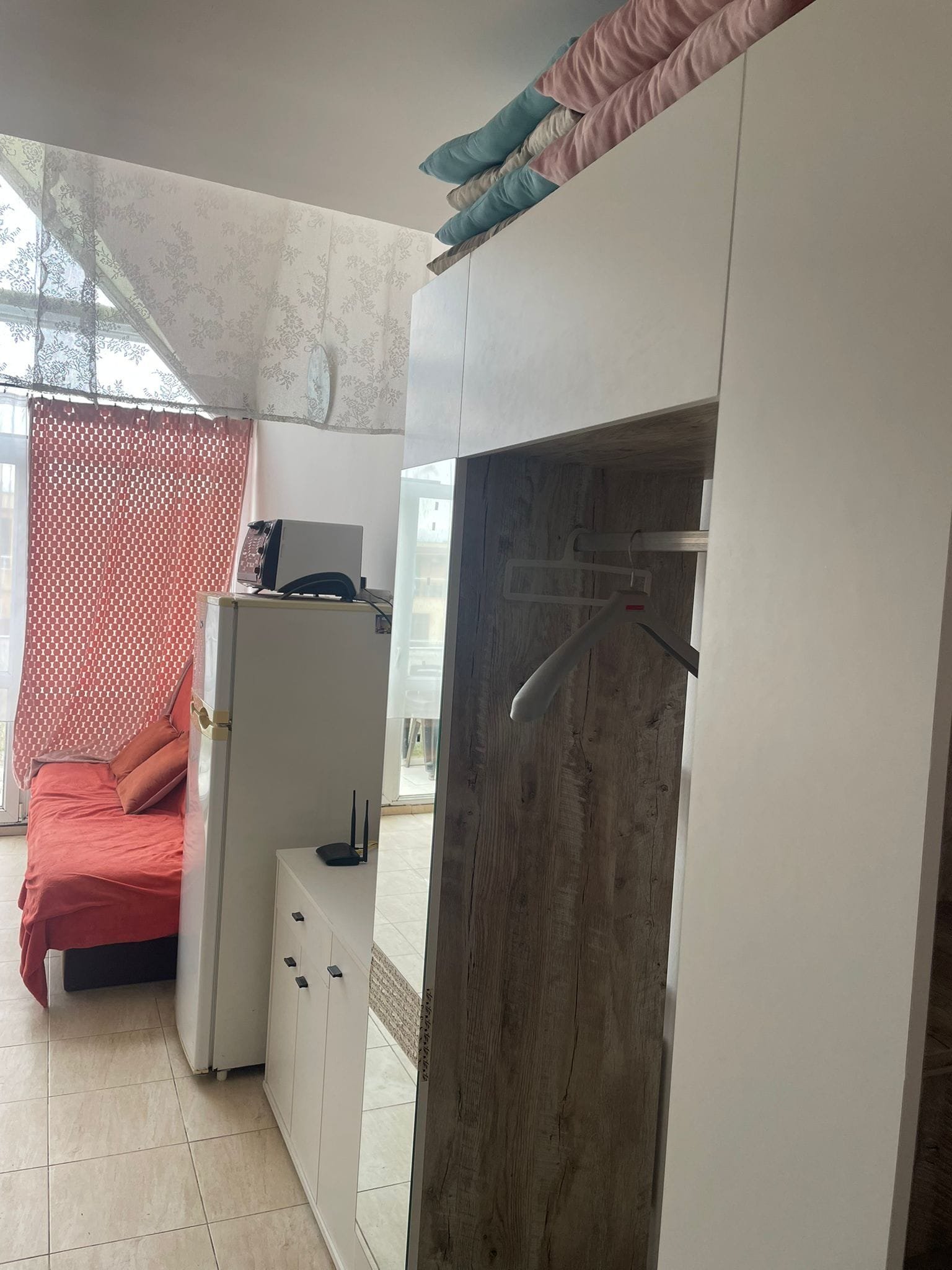 Студия с жилой зоной, диваном, холодильником и шкафом, оформленная в светлых тонах с акцентами на текстиле.