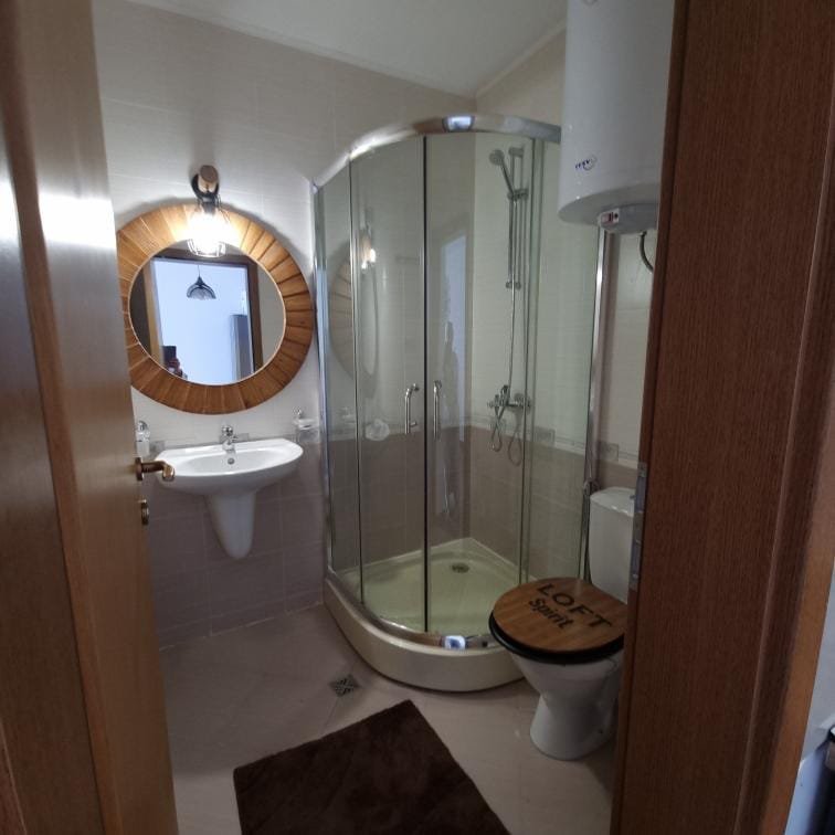 Современная ванная комната с душевой кабиной, туалетом, раковиной и круглым зеркалом в деревянной рамке.