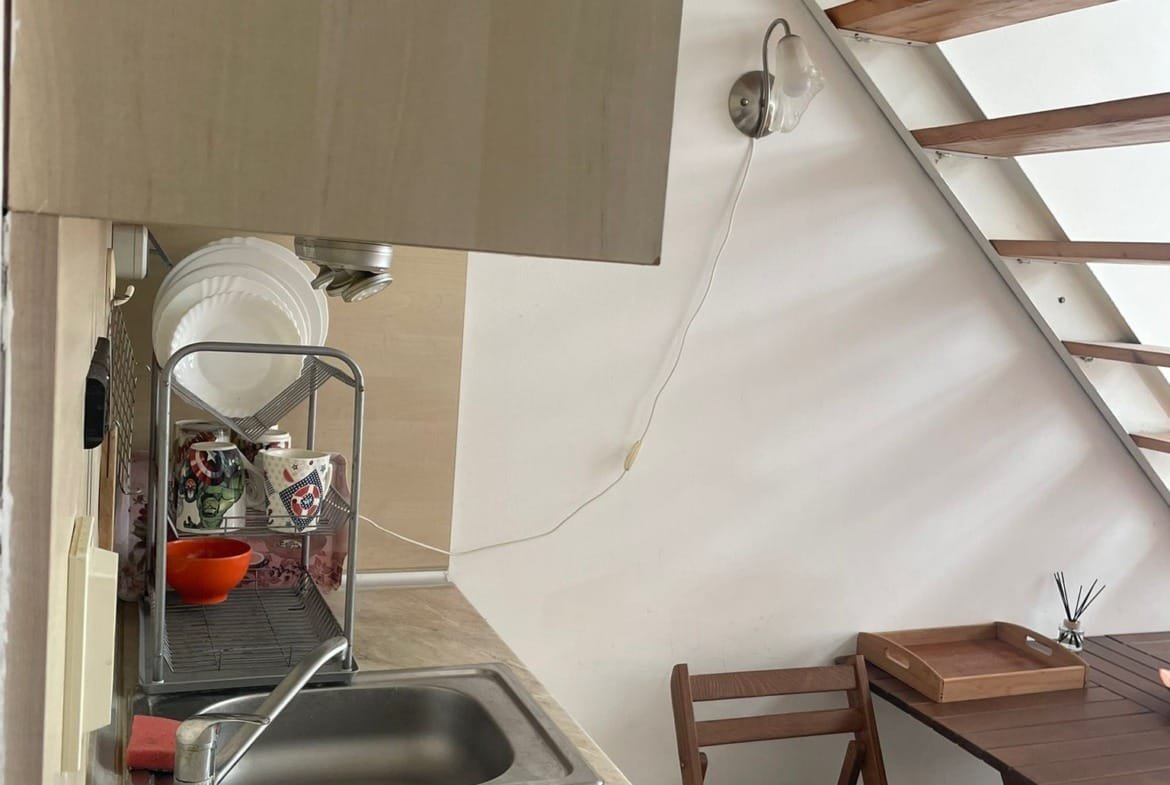 Кухонный уголок в студии с мойкой, деревянной лестницей и столом с предметами быта. Классический дизайн, уютная атмосфера.