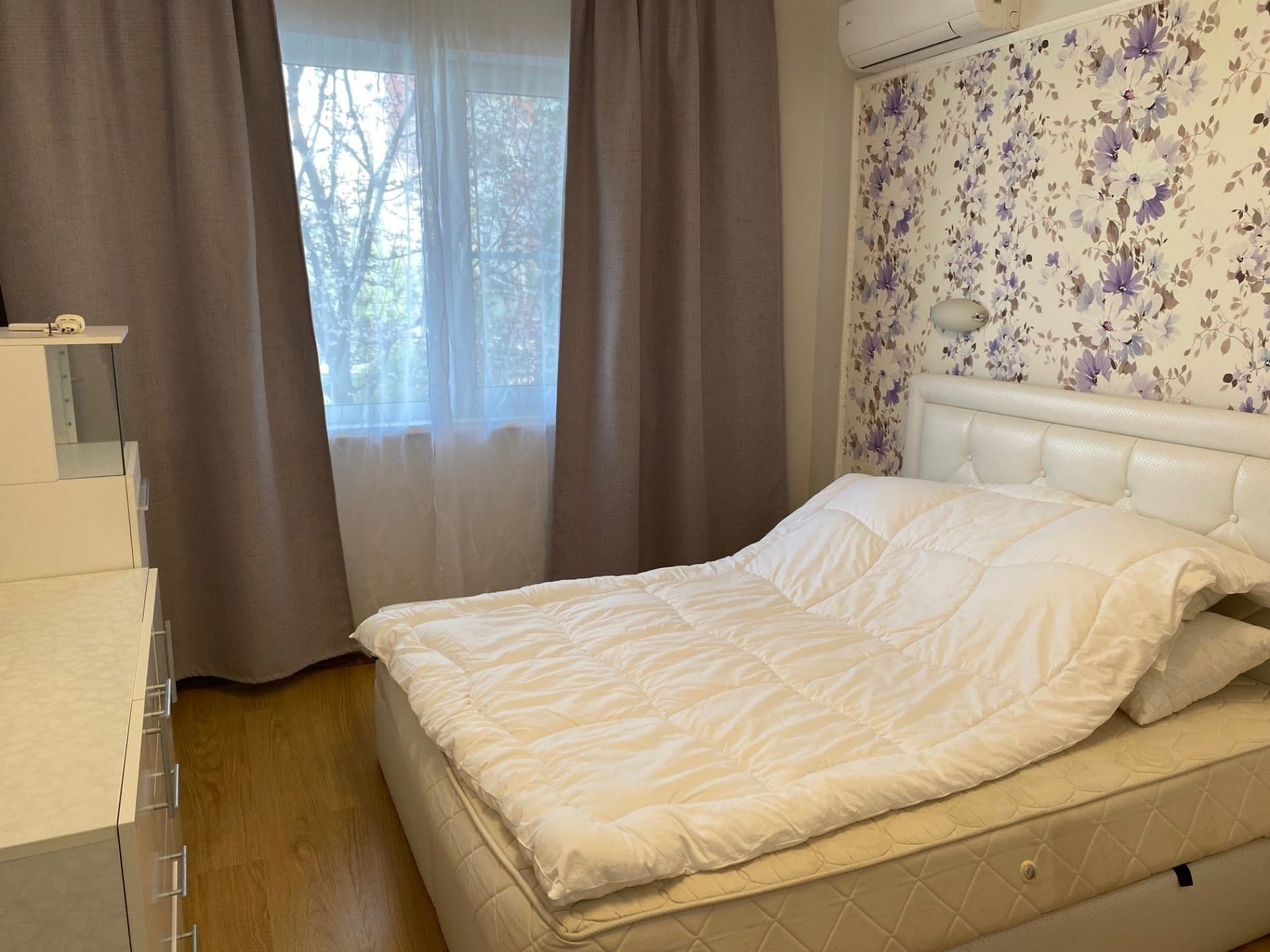 Спальня в трехкомнатном апартаменте с двуспальной кроватью, светлыми шторами и цветочной обоями, окно с видом на природу.