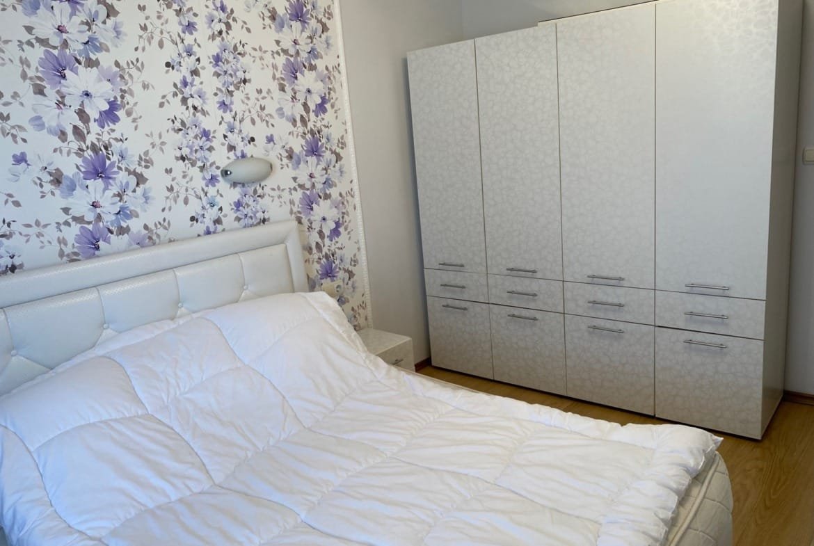 Спальня с белой кроватью и шкафом, оформленная фиолетовыми цветами на стене, создающая уютную атмосферу.