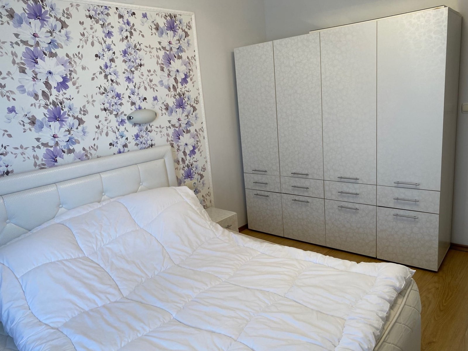 Спальня с белой кроватью и шкафом, оформленная фиолетовыми цветами на стене, создающая уютную атмосферу.