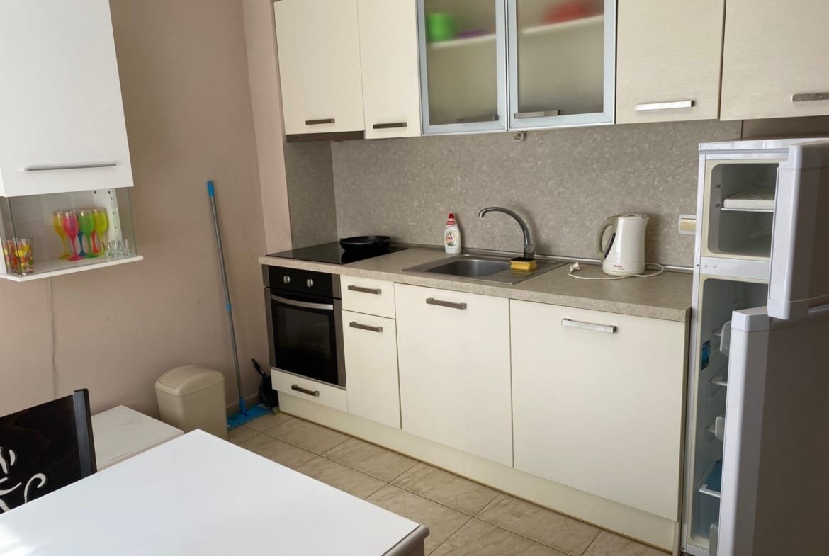 Современная кухня с белыми фасадами, плитой, раковиной, холодильником и обеденным столом. Просторная и светлая планировка.