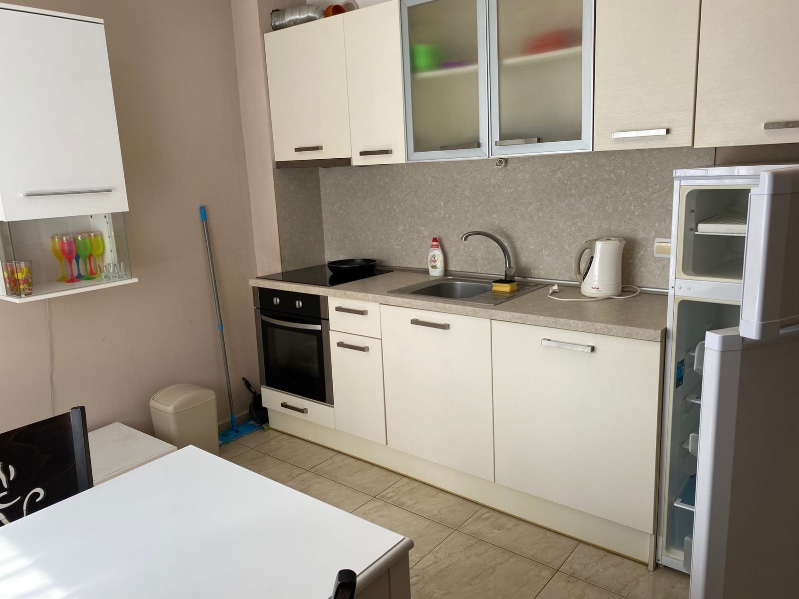 Современная кухня с белыми фасадами, плитой, раковиной, холодильником и обеденным столом. Просторная и светлая планировка.