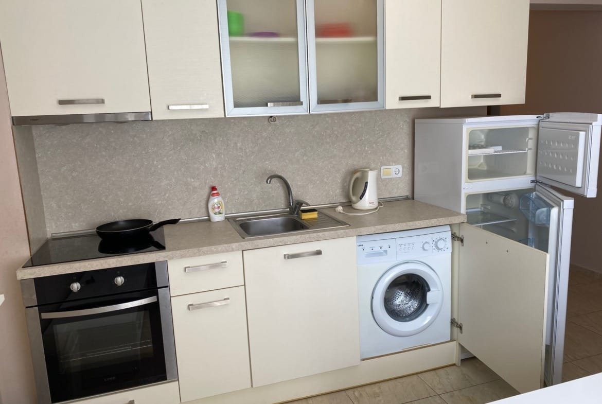 Современная кухня с белыми шкафчиками, встроенной техникой, холодильником, стиральной машиной и мойкой.