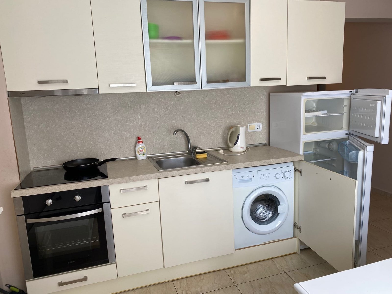 Современная кухня с белыми шкафчиками, встроенной техникой, холодильником, стиральной машиной и мойкой.