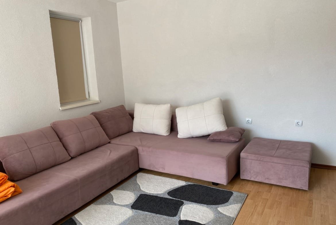 Современный угловой диван пурпурного цвета с подушками, рядом серый ковёр и светлые стены в трехкомнатном апартаменте.