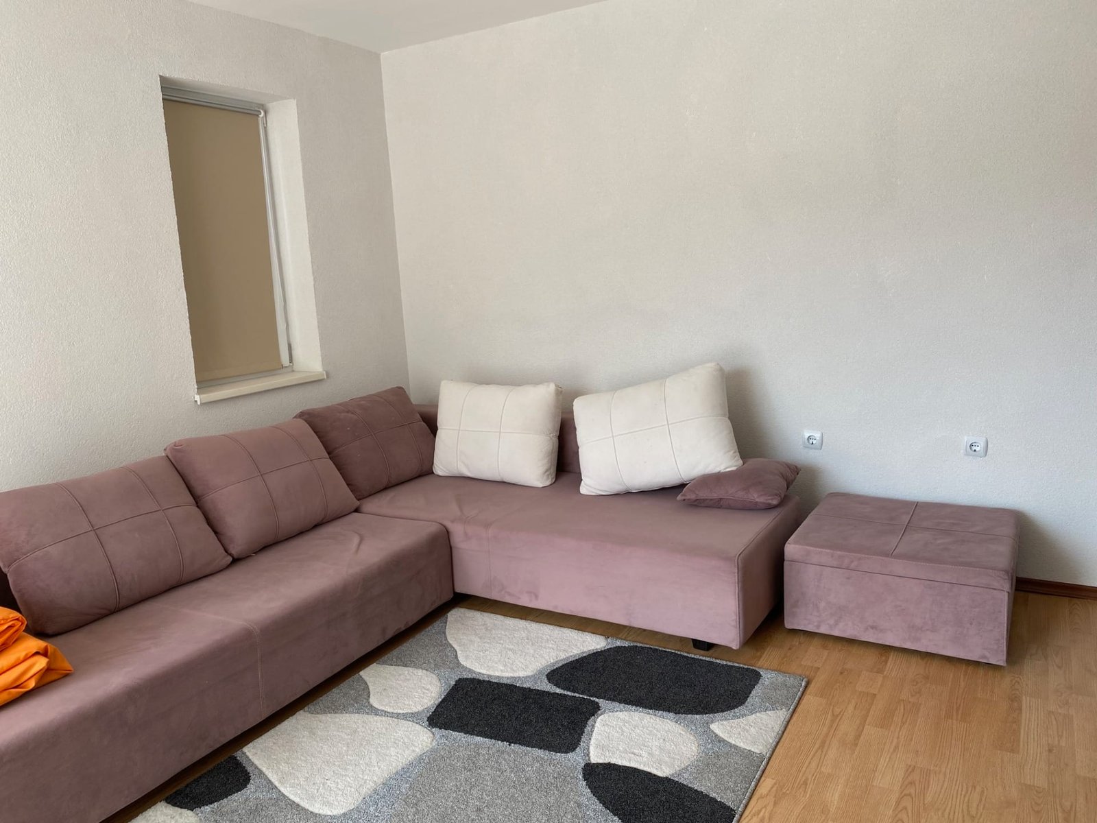 Современный угловой диван пурпурного цвета с подушками, рядом серый ковёр и светлые стены в трехкомнатном апартаменте.