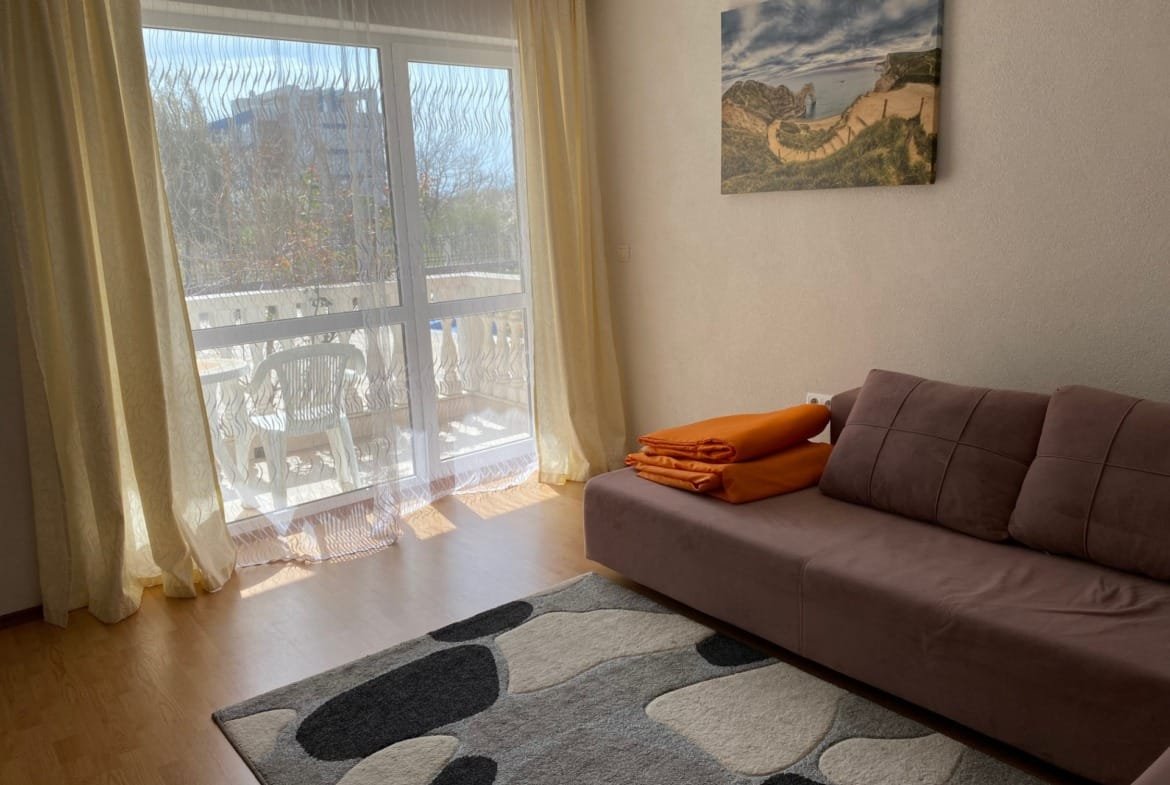 Современная гостиная с мягким диваном, оранжевыми пледами и большими окнами, выходящими на балкон.