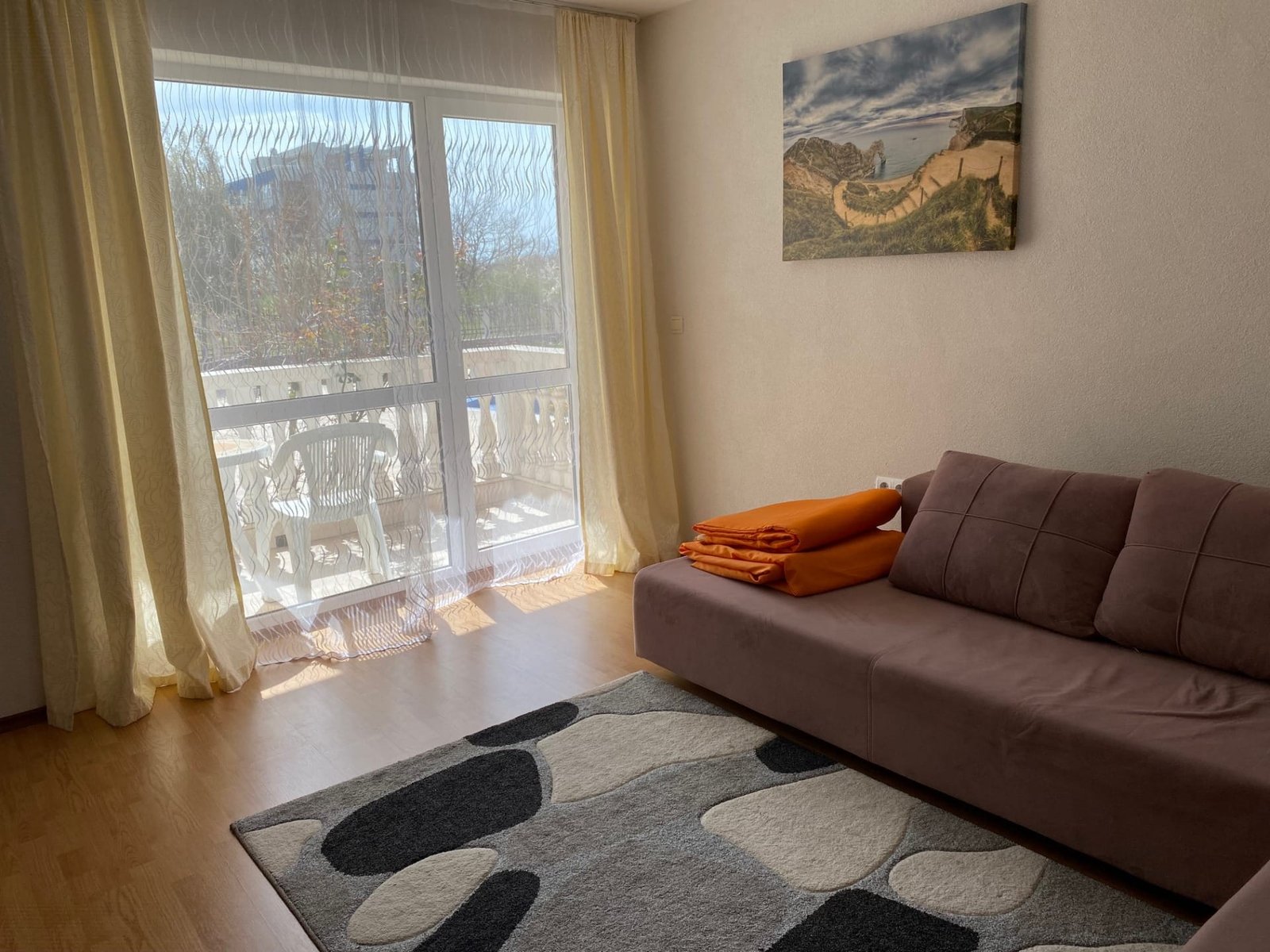 Современная гостиная с мягким диваном, оранжевыми пледами и большими окнами, выходящими на балкон.