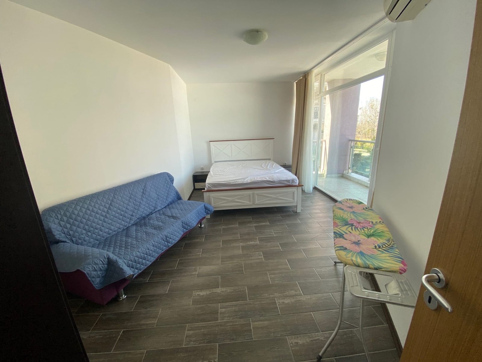 Современная комната с односпальной кроватью, диваном и гладильной доской, выход на балкон с видом на окружающую природу.