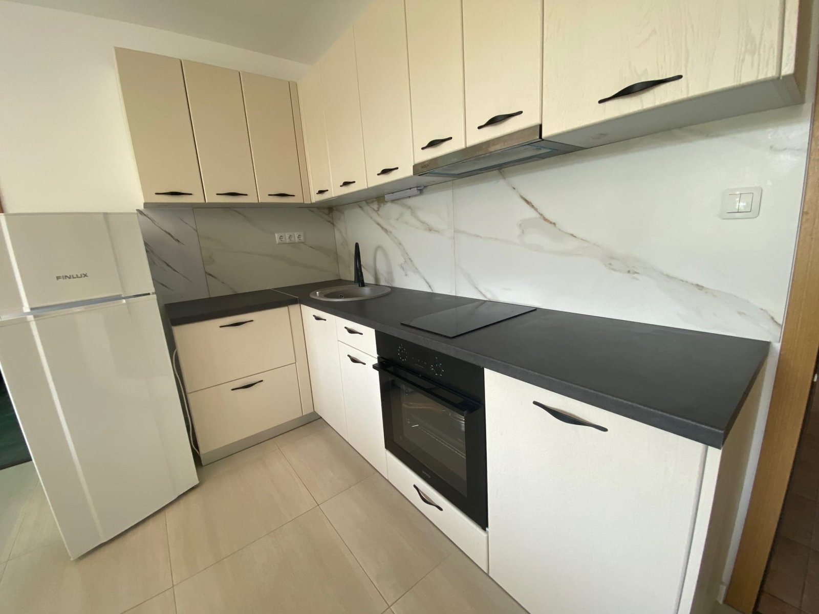 Современная кухня в трёхкомнатном апартаменте с белыми и серыми шкафами, чёрной столешницей и бытовой техникой.