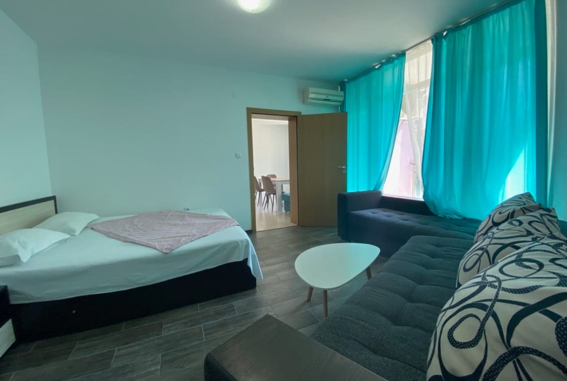 Современный интерьер трехкомнатного апартамента: кровать, темный диван, светлые занавески, столик и дверь в кухню.