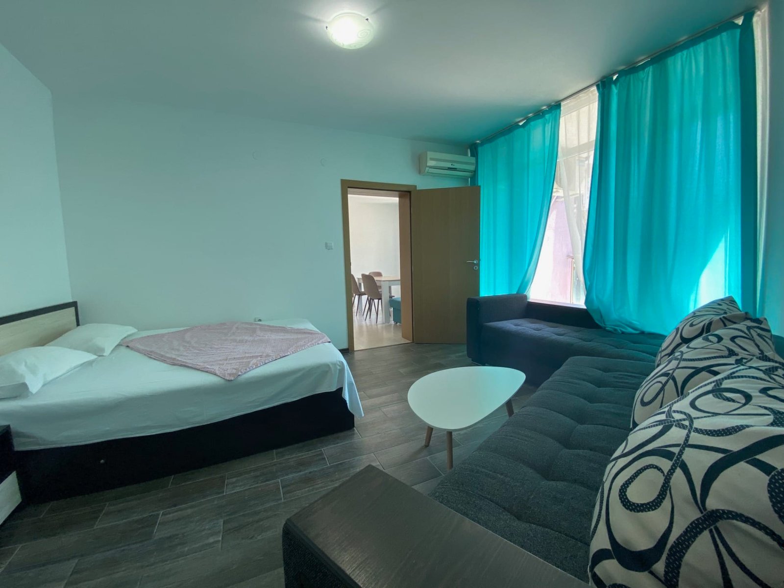 Современный интерьер трехкомнатного апартамента: кровать, темный диван, светлые занавески, столик и дверь в кухню.