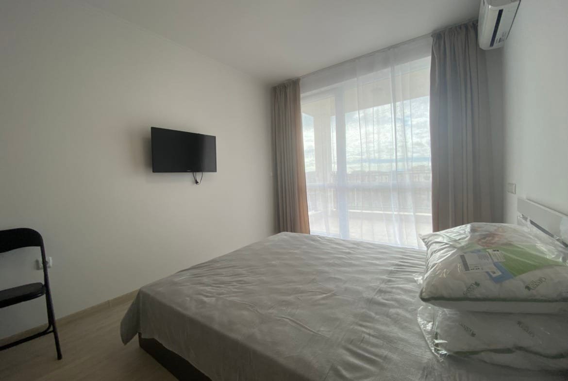 Современная студия в комплексе с кроватью, телевизором и панорамными окнами, оформленная в светлых тонах.