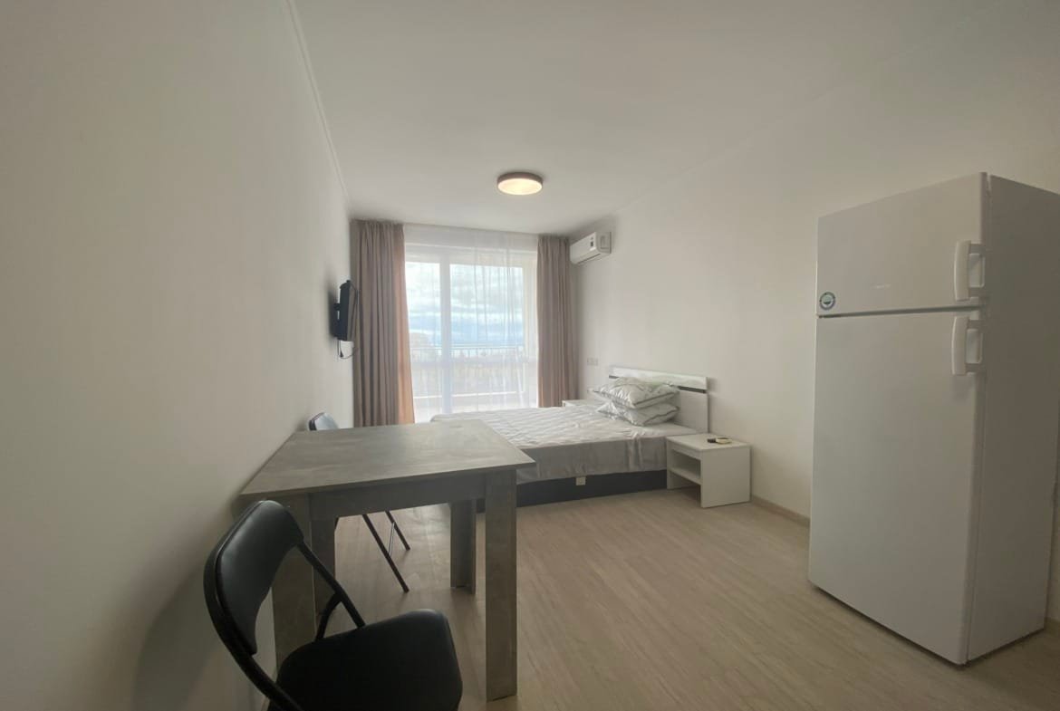 Современная студия с холодильником, столом и стульями, светлые стен и окна с видом на улицу.