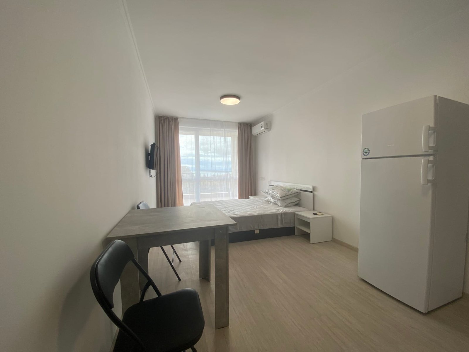 Современная студия с холодильником, столом и стульями, светлые стен и окна с видом на улицу.